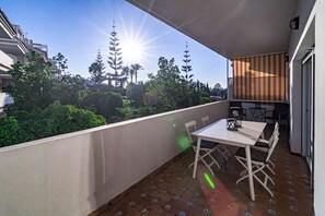 Un balcón con mesa y sillas, vista a árboles y edificios, y un sol brillando al fondo.