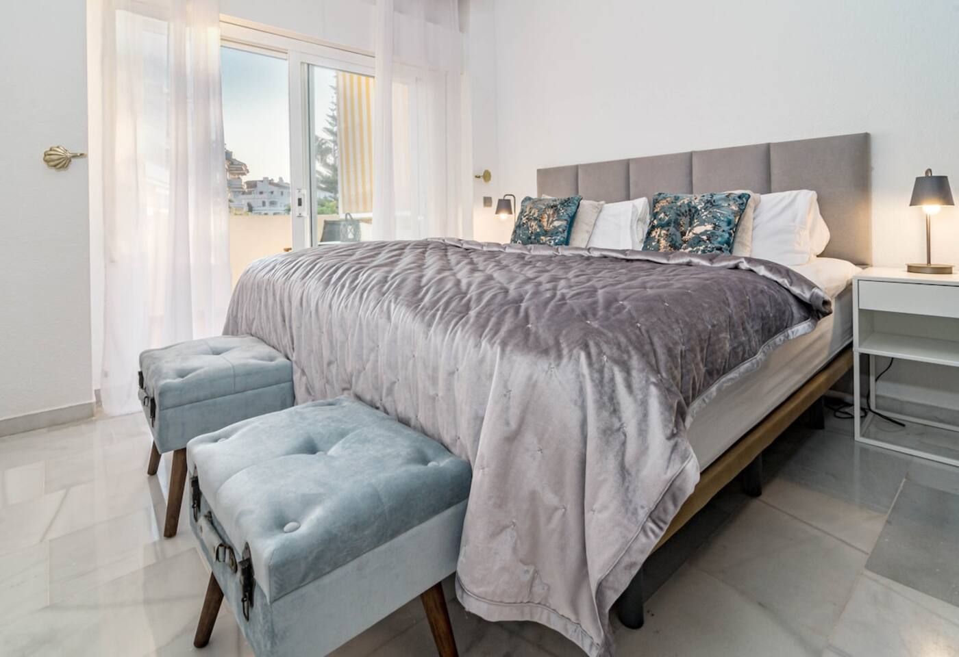 A modern bedroom with a large bed, a bench, a nightstand, and a view of the outside.