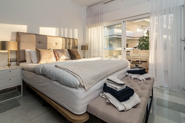 Ein modernes Schlafzimmer mit einem großen Bett, Nachttischlampen, einer Bank mit Handtüchern und Blick auf den Balkon.