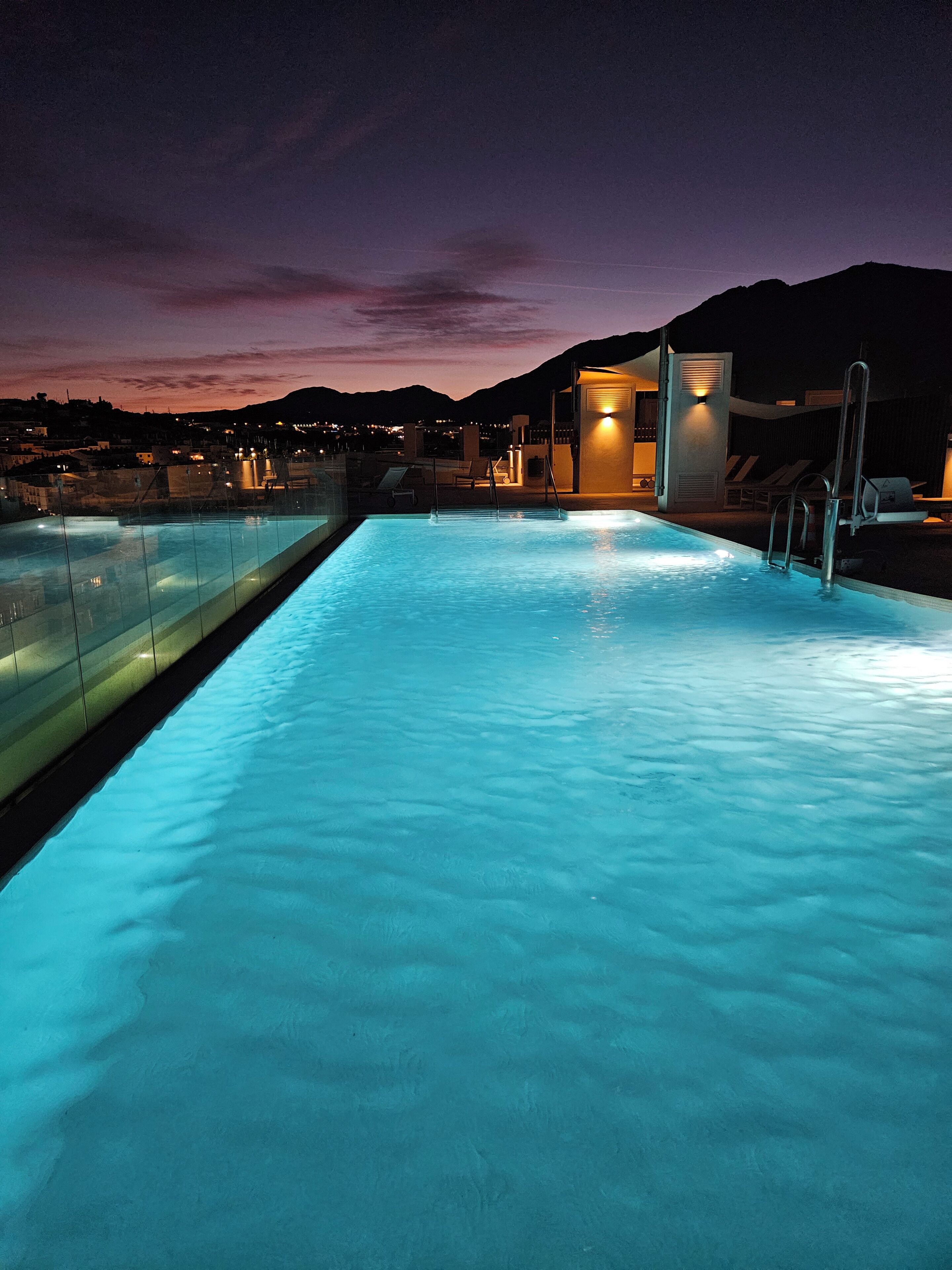 Une piscine à l’eau claire, une barrière en verre et un petit bâtiment éclairé.