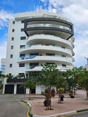 Un edificio de varios pisos con balcones, fachada curva y una zona de estacionamiento con plantas en macetas.