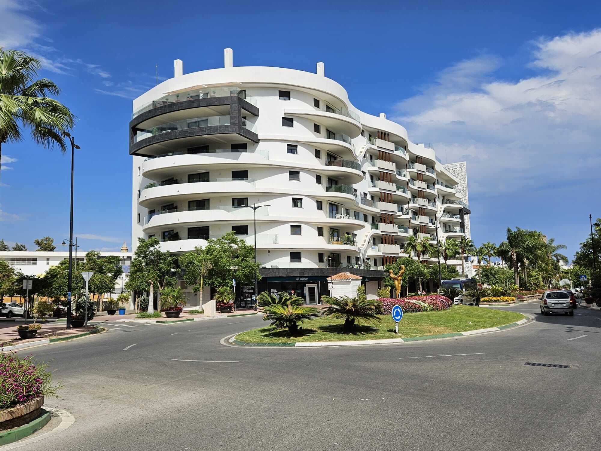 A multi-story hotel with a curved facade, balconies, and a landscaped area with palm trees.