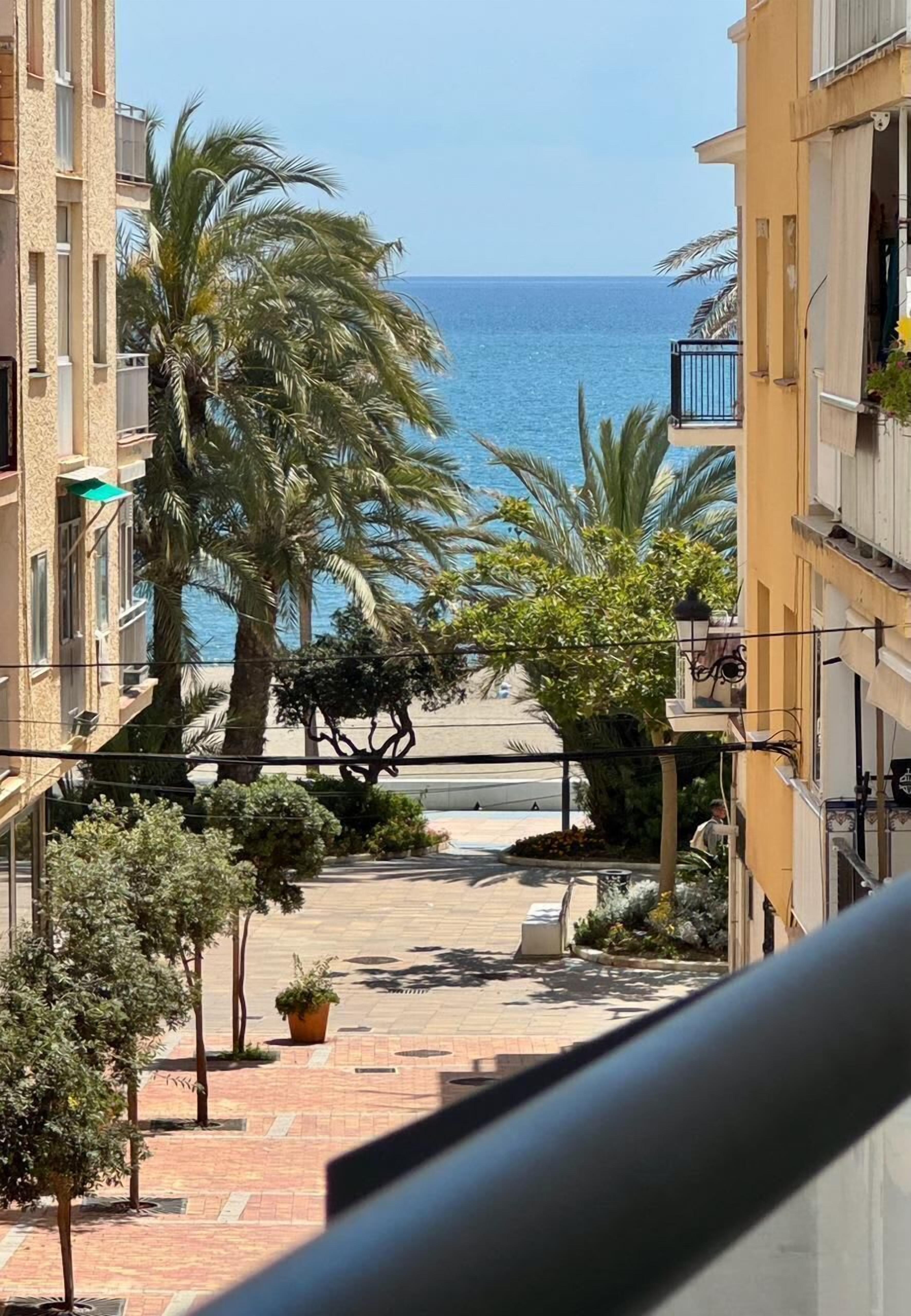 A residential building with a view of the sea, palm trees, and a paved pathway.
