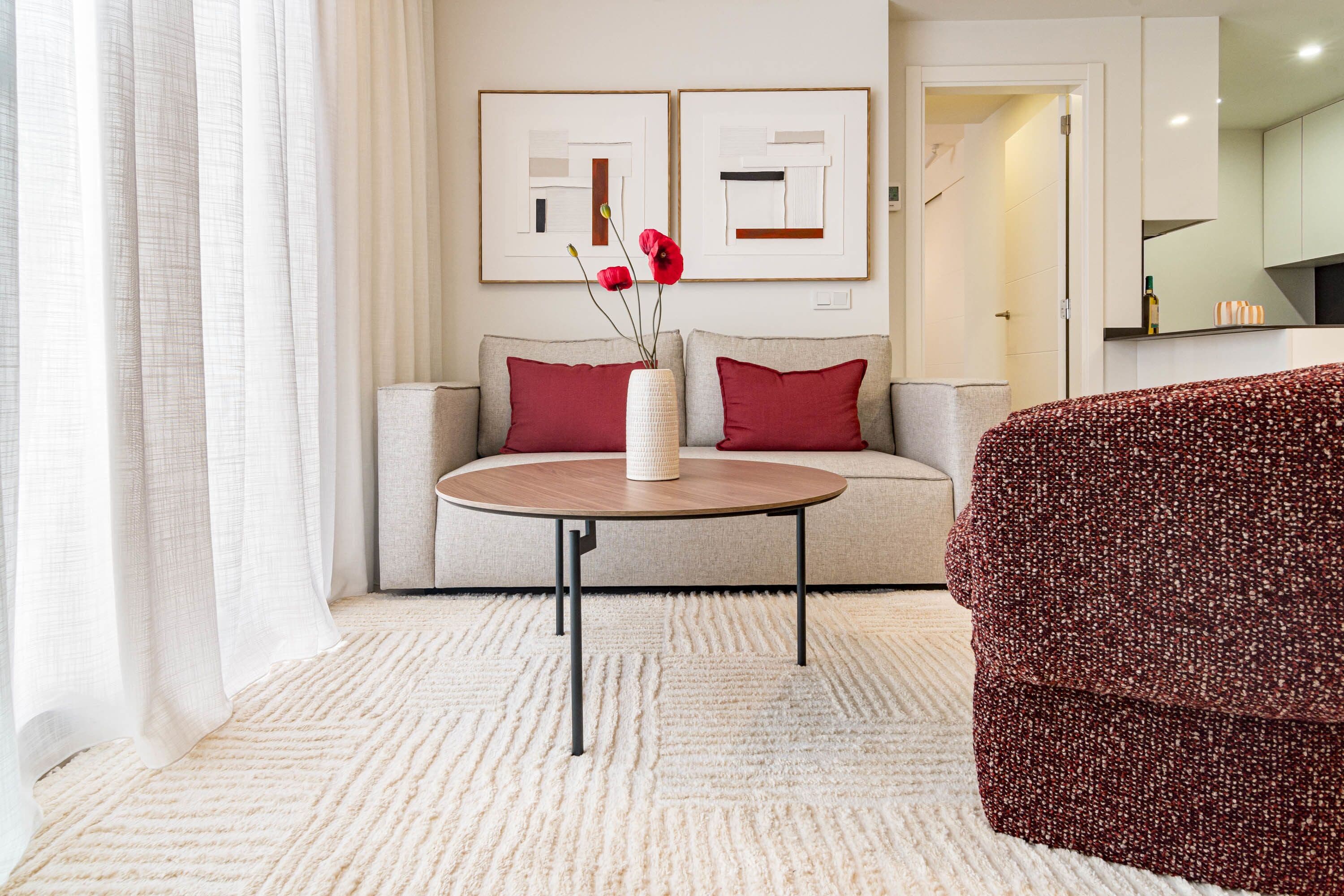 A living room with a sofa, a round coffee table, and a red armchair. There are two framed abstract paintings on the wall.