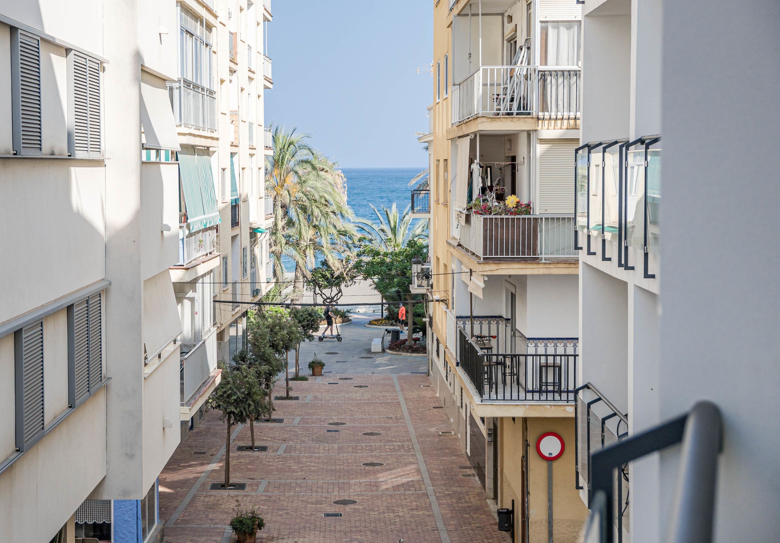 Une rue étroite en milieu urbain, bordée d’immeubles, avec un ciel dégagé et une vue lointaine sur la mer.