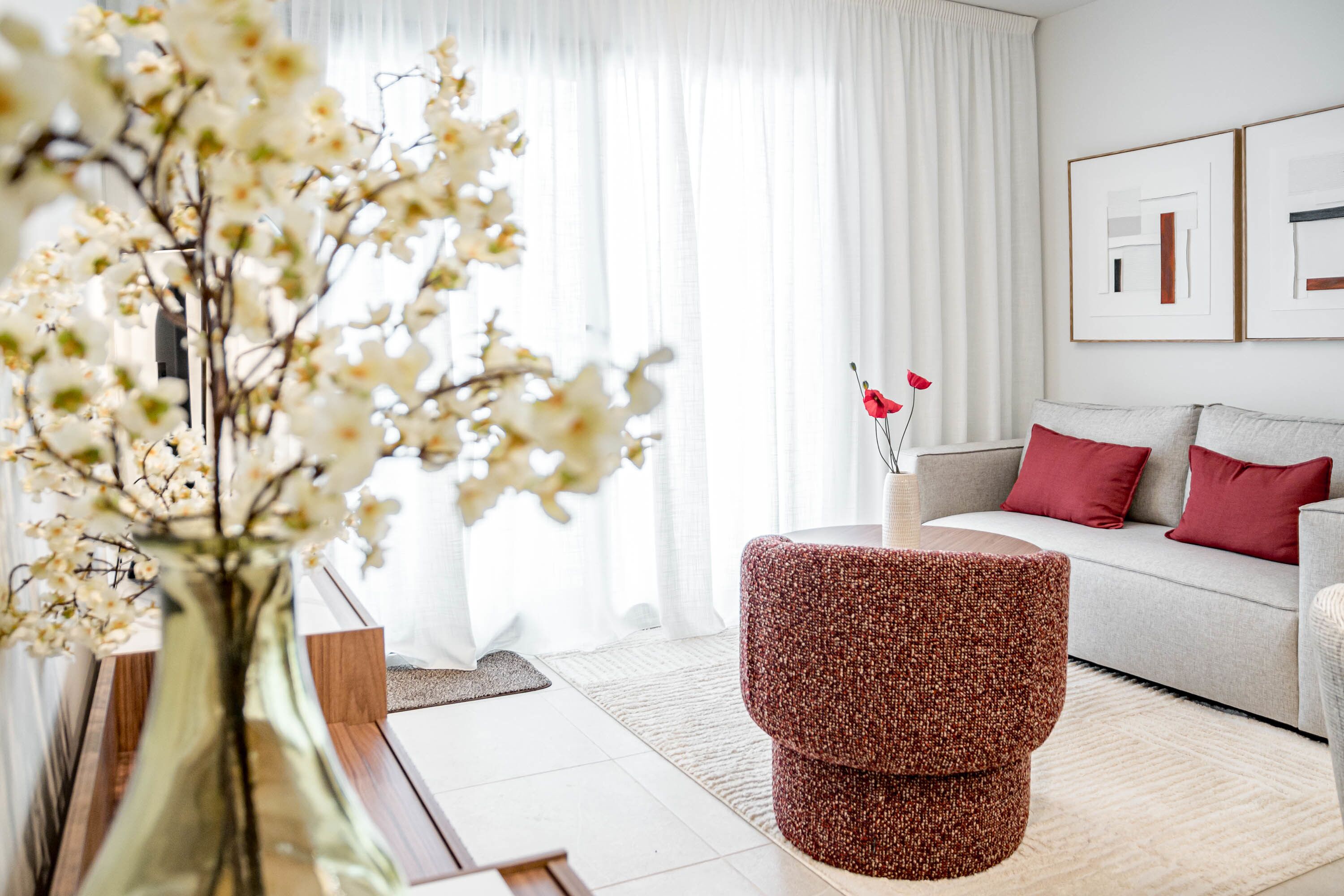 A modern living room with a sofa, a red accent stool, a vase with flowers, and framed artwork on the wall.