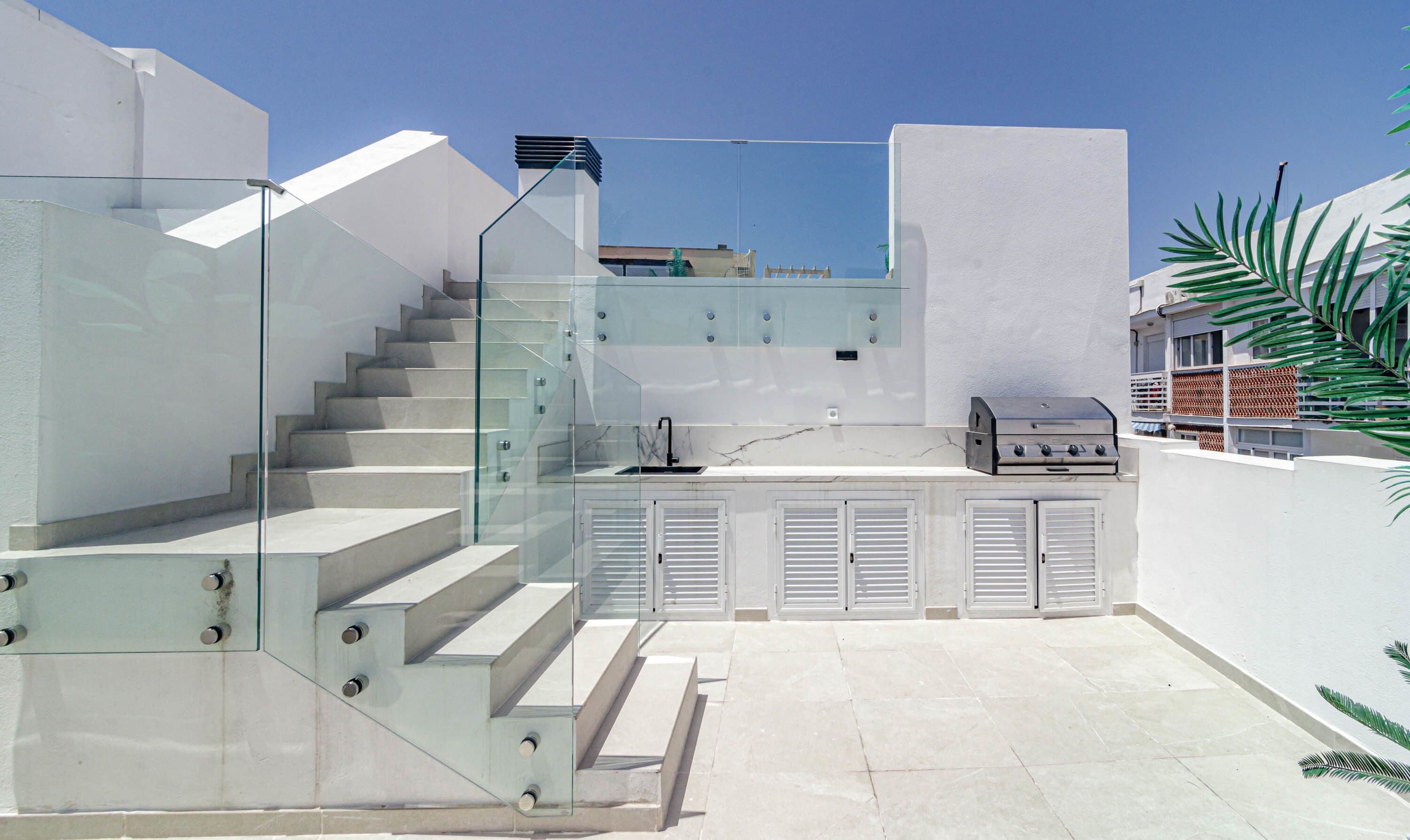 Une cuisine extérieure moderne, dotée d’un escalier en verre, d’un barbecue et offrant une vue sur le ciel.