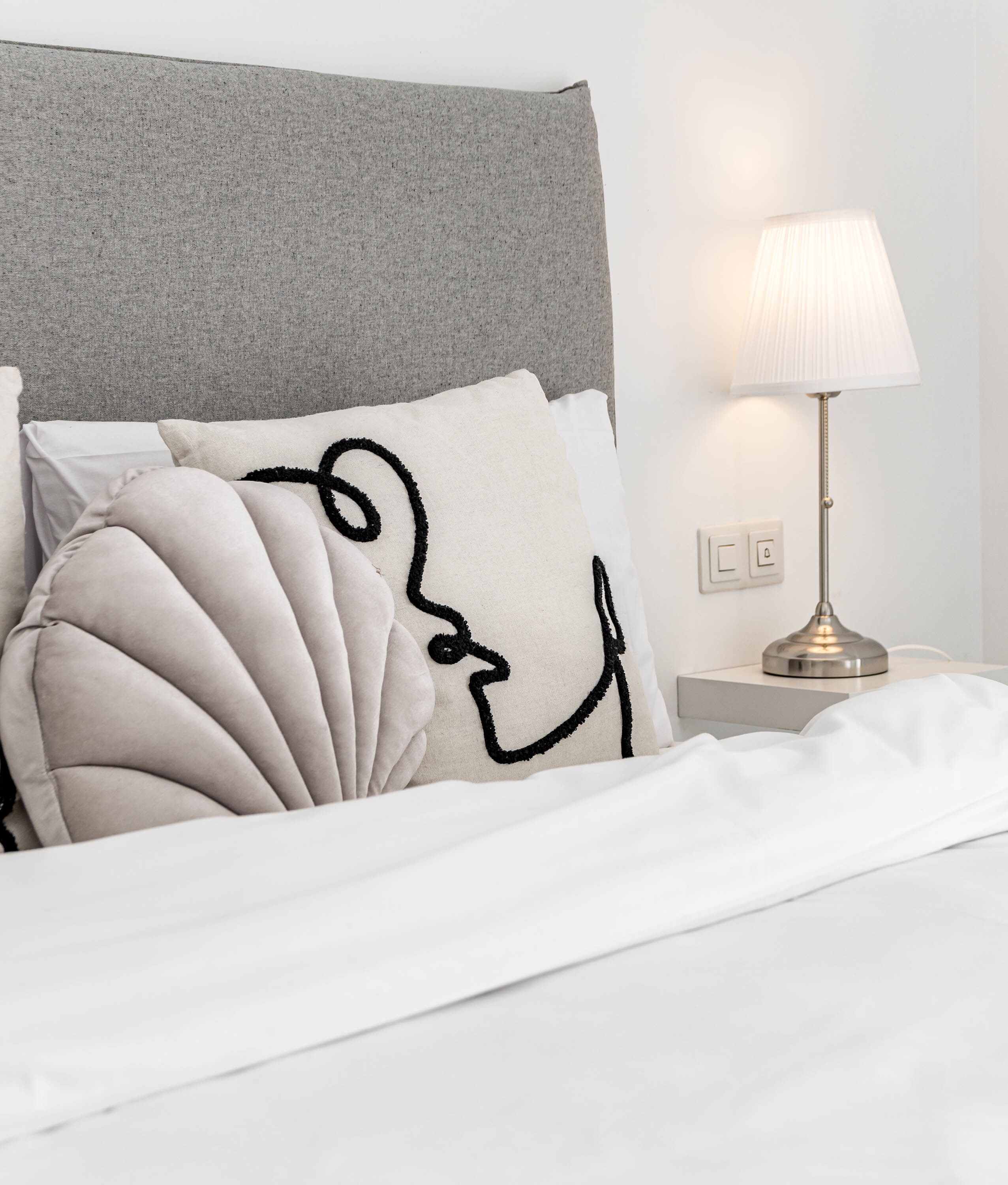 A neatly made bed with a grey headboard, white bedding, and decorative pillows including one with a black line drawing. A bedside lamp is on to the right.