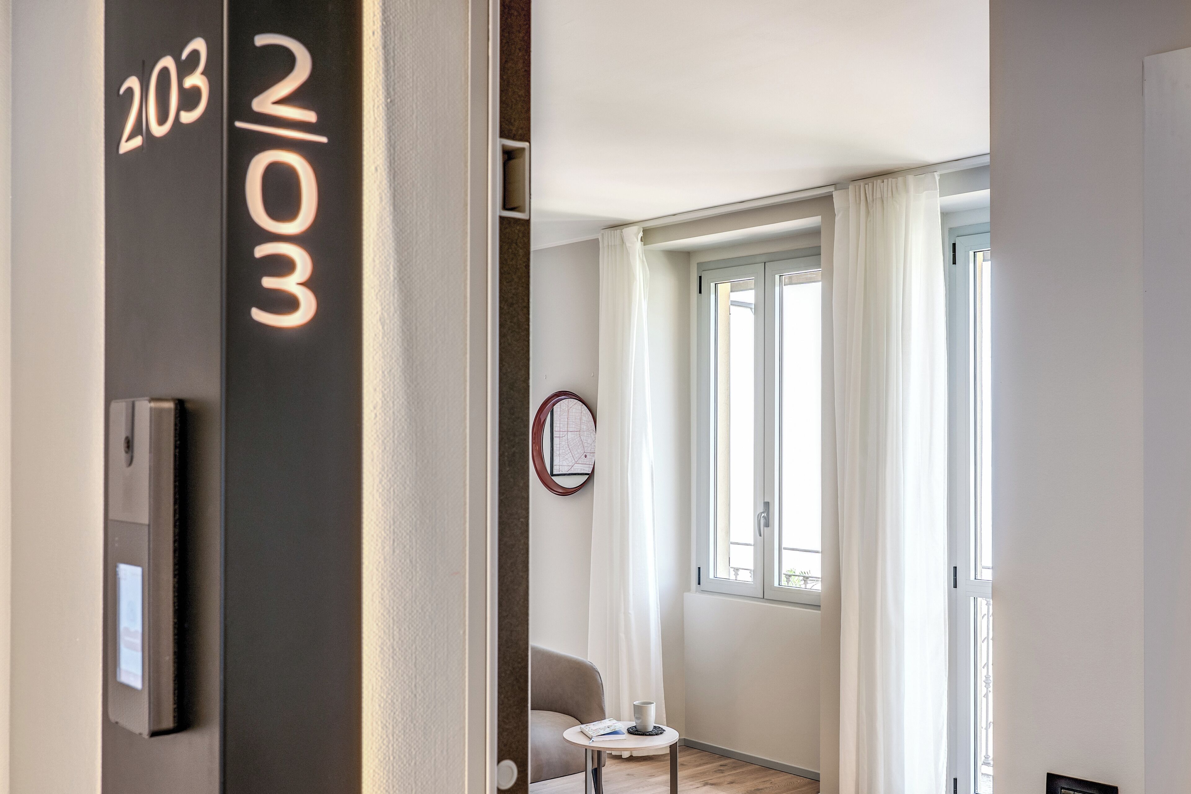 Une chambre d’hôtel moderne avec une grande fenêtre, une petite table sur laquelle se trouve une tasse, et une carte accrochée au mur.