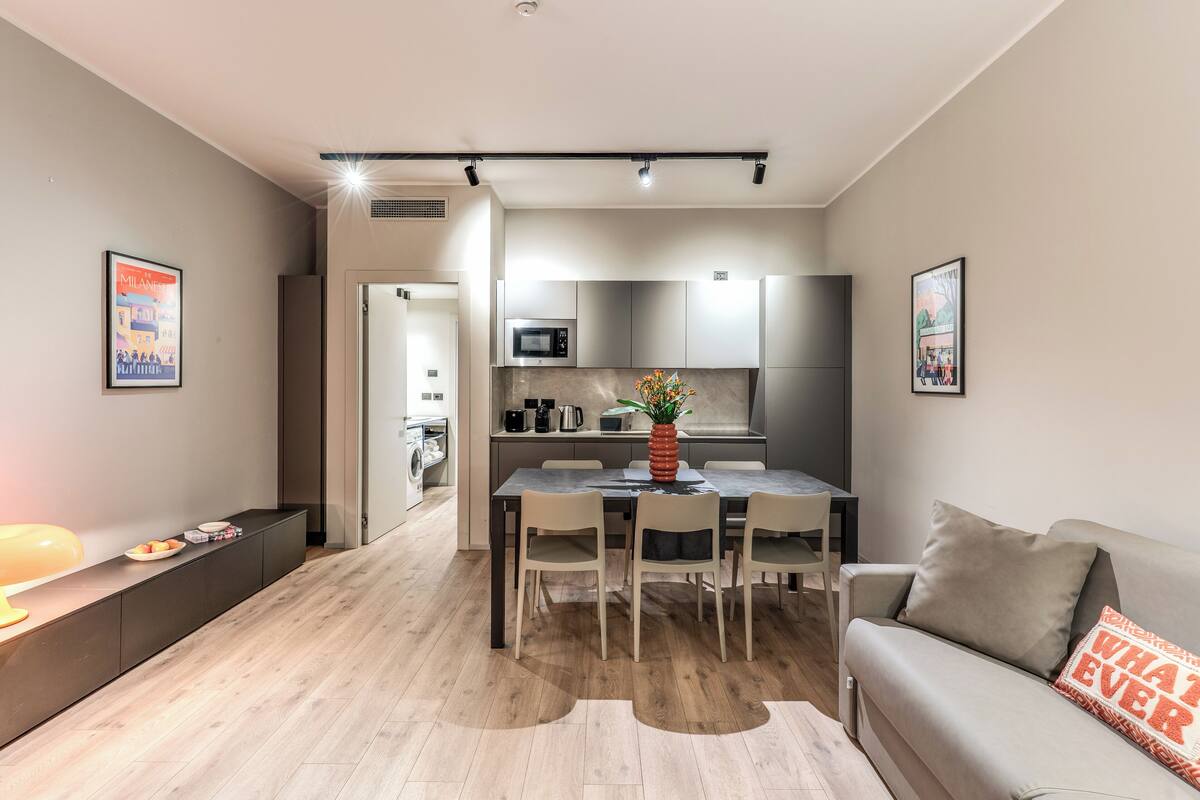 A modern living room with a dining area, a kitchen in the background, and a sofa with a cushion.