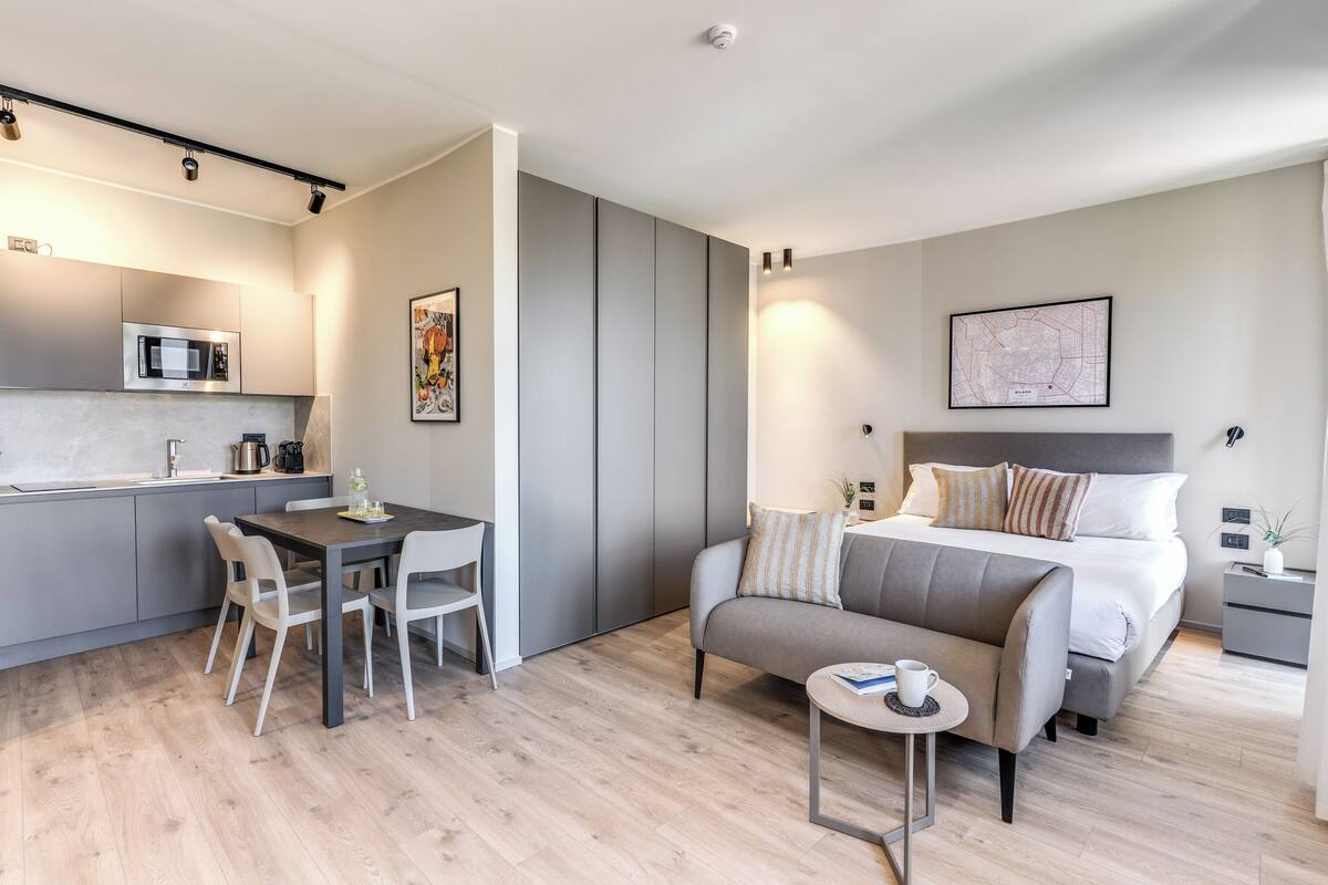 A modern hotel room with a kitchenette, a bed with multiple pillows, a sofa, a small table, and a coffee machine.