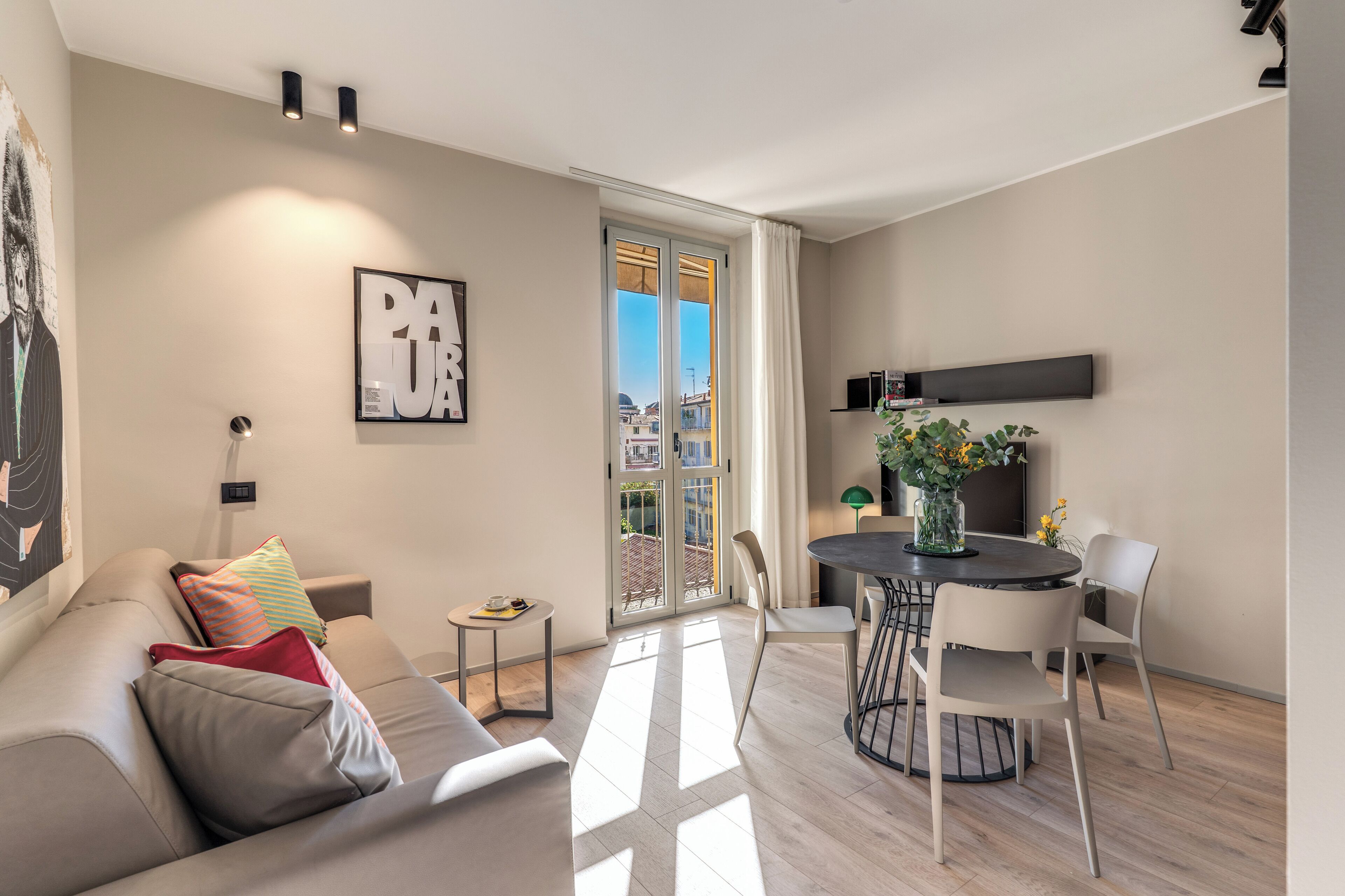 A modern living room with a sofa, a dining table with chairs, a small side table, and a large window with a view of buildings.