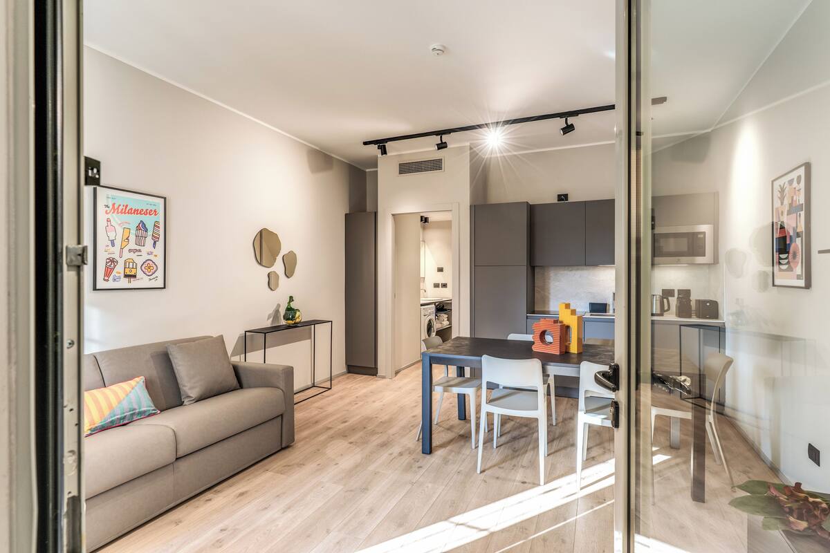 A modern living room with a sofa, a dining table, and chairs. There is a kitchen area with cabinets and appliances visible in the background.