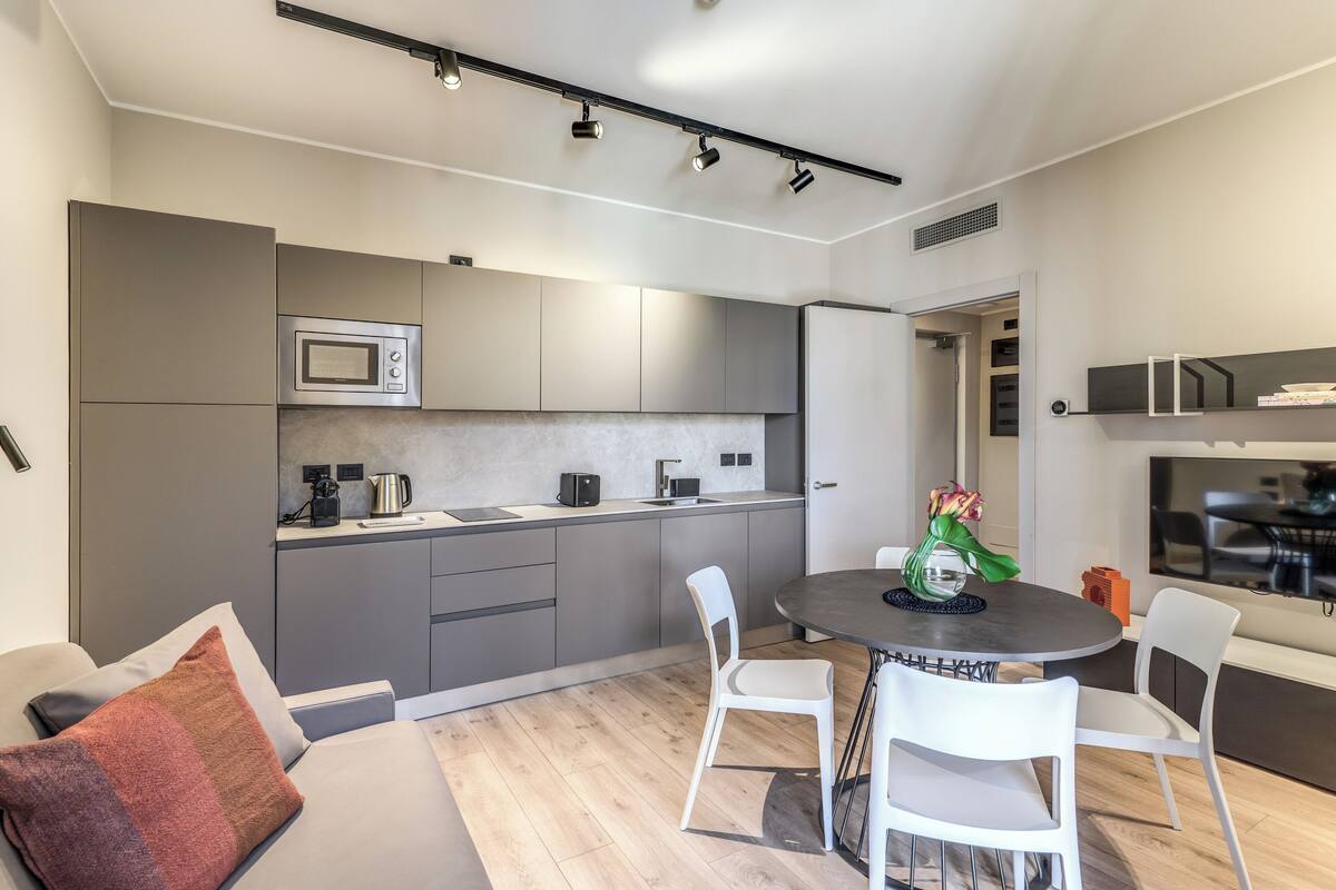 A modern kitchen with built-in appliances, a dining area with a round table and white chairs, and a cozy seating area with a sofa and cushions.