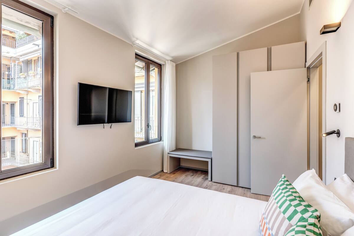 A bedroom with a bed, a TV mounted on the wall, a bench, a wardrobe, and a window with a view of buildings.