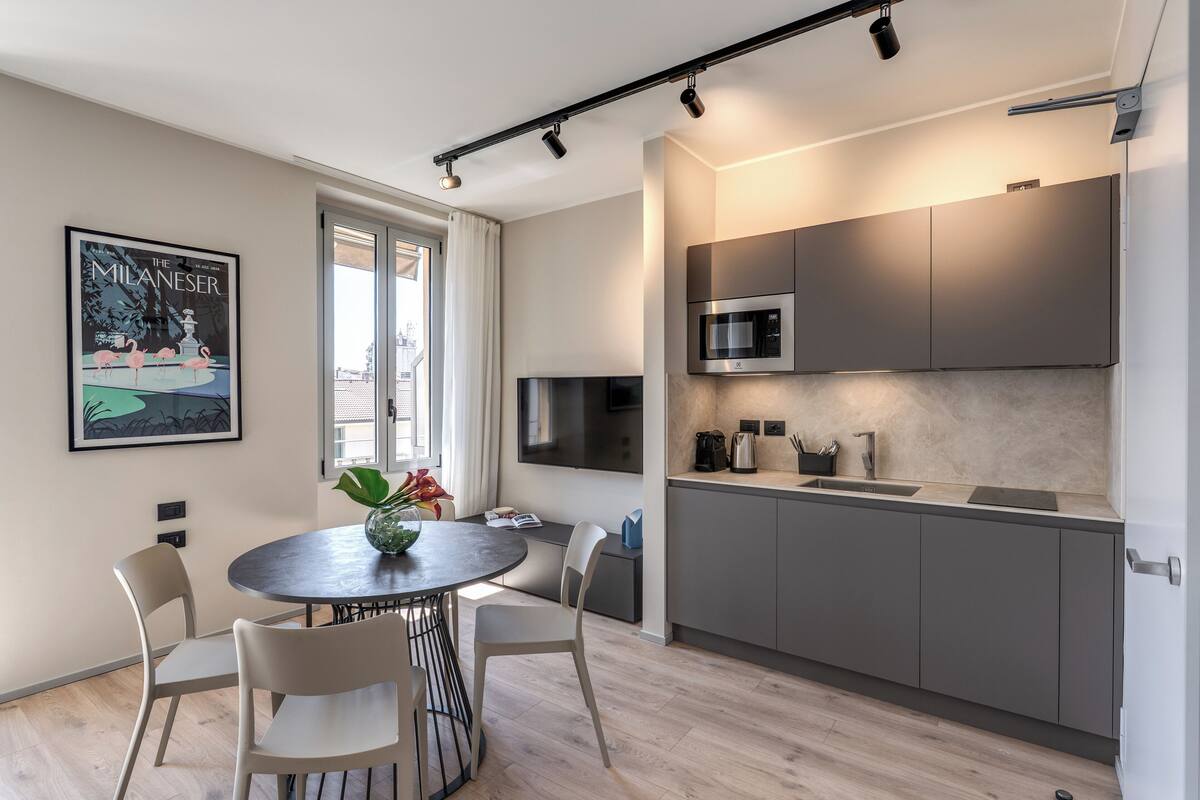 A modern kitchen with grey cabinets, a built-in microwave, and a coffee machine. There is a dining area with a round table and chairs, and a window providing natural light.