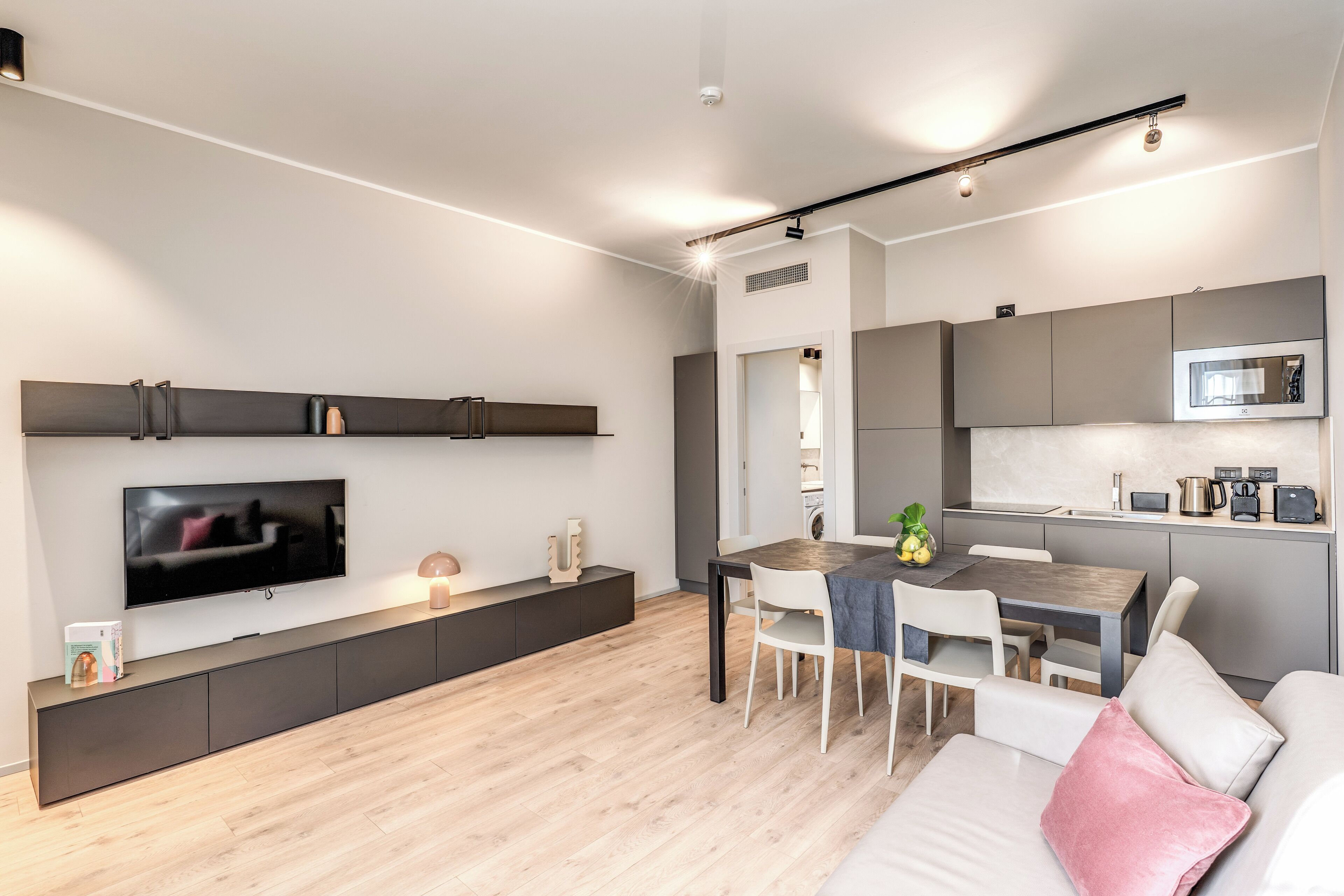 A modern living room with a flat-screen TV, a dining area with a wooden table and chairs, and a kitchen with grey cabinets and a microwave.