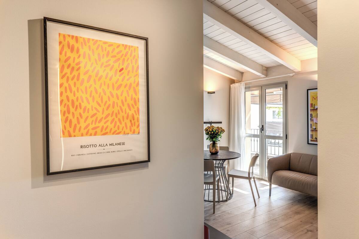 A framed artwork titled "RISOTTO ALLA MILANESI" on a wall, a dining area with a table and chairs, and a sofa in a well-lit room with a wooden ceiling.