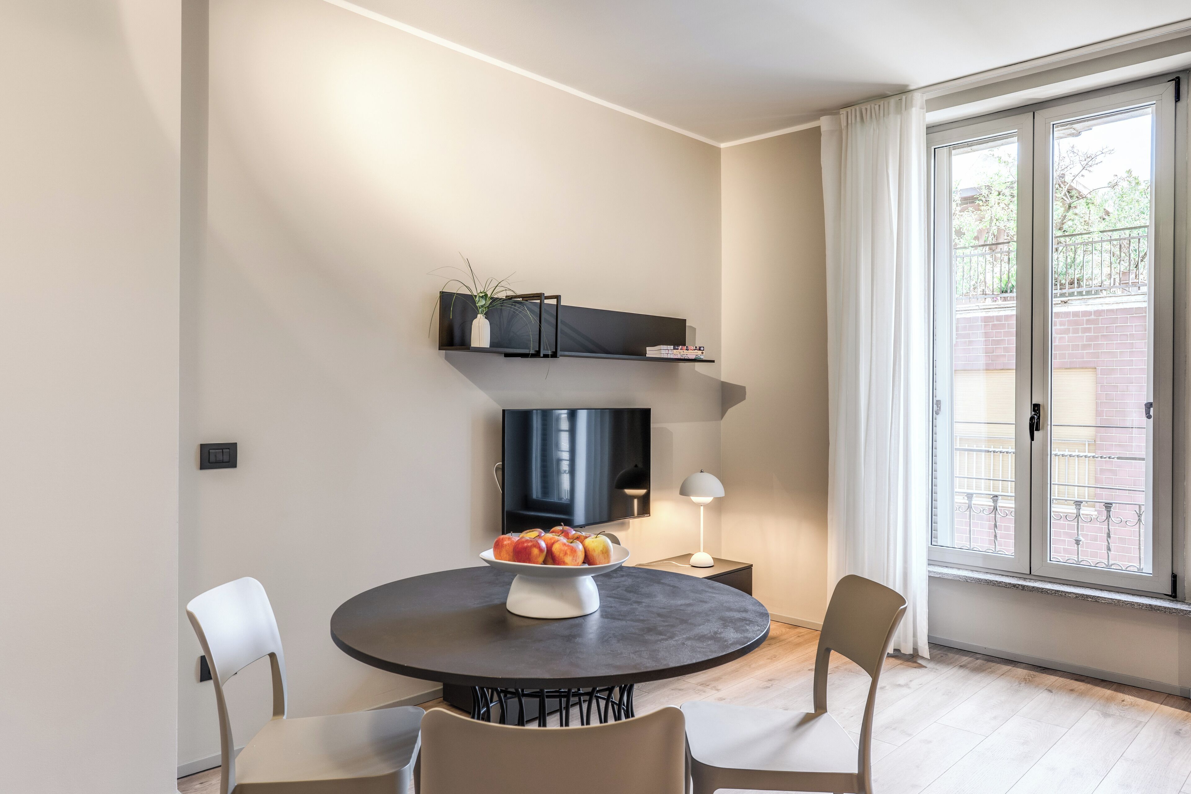 A modern dining area with a round table, chairs, a bowl of fruit, a TV, and a window with curtains.