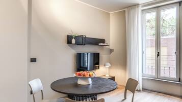 A modern dining area with a round table, chairs, a bowl of fruit, a TV, and a window with curtains.