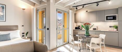 A modern hotel room with a bed, a sofa, a dining table with chairs, a kitchen area, and a balcony with a view.