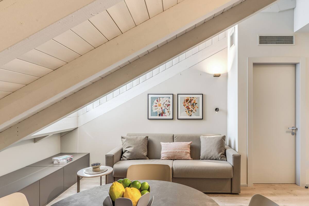 A modern living room with a high ceiling, a built-in bench, a round table with fruit, a sofa with cushions, and two framed artworks on the wall.