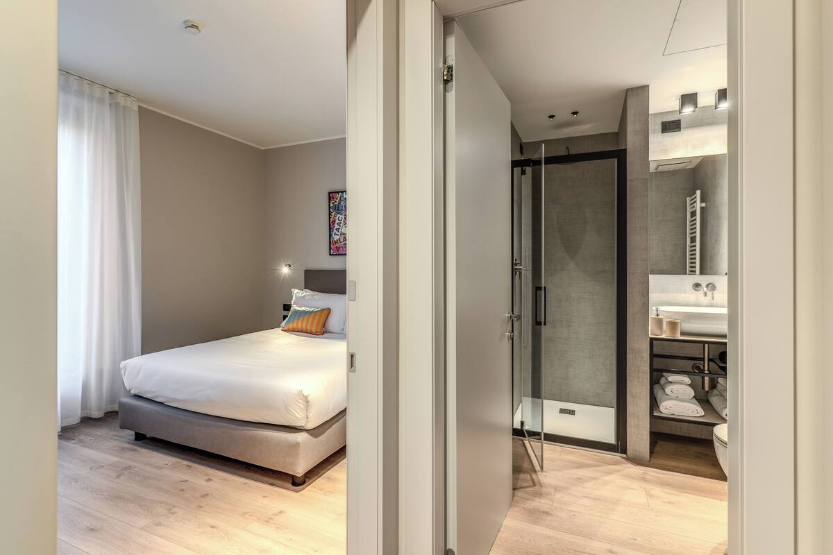 A modern hotel room with a bed, a shower, and a bathroom with a sink and towel rack.
