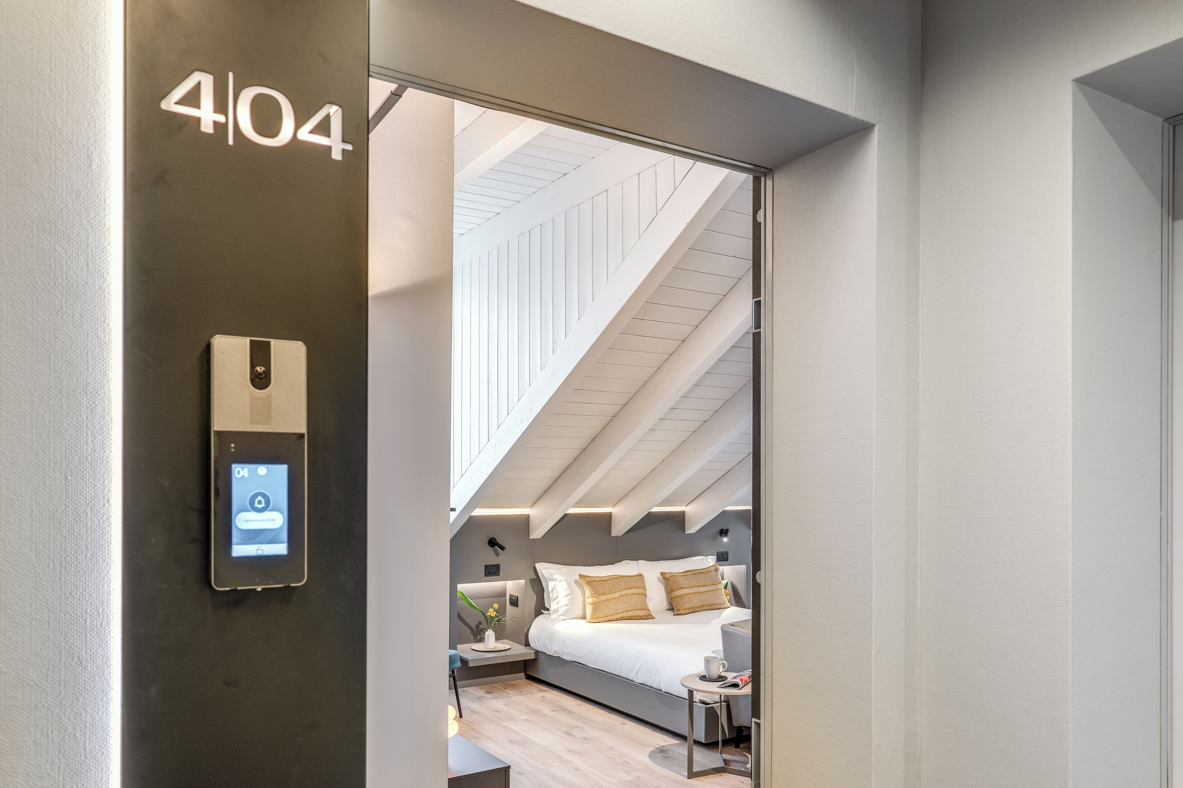 A modern hotel room with a digital lock, a bed with white bedding, a bedside table with a lamp, and a view of the attic.