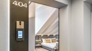 A modern hotel room with a digital lock, a bed with white bedding, a bedside table with a lamp, and a view of the attic.