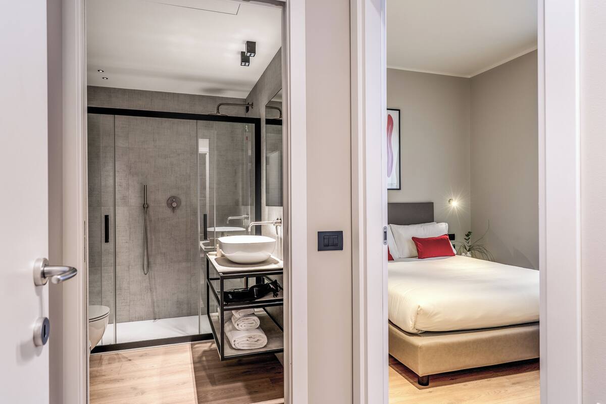 A modern hotel room with a glass shower enclosure, a white sink, a bedside table, and a bed with a red pillow.
