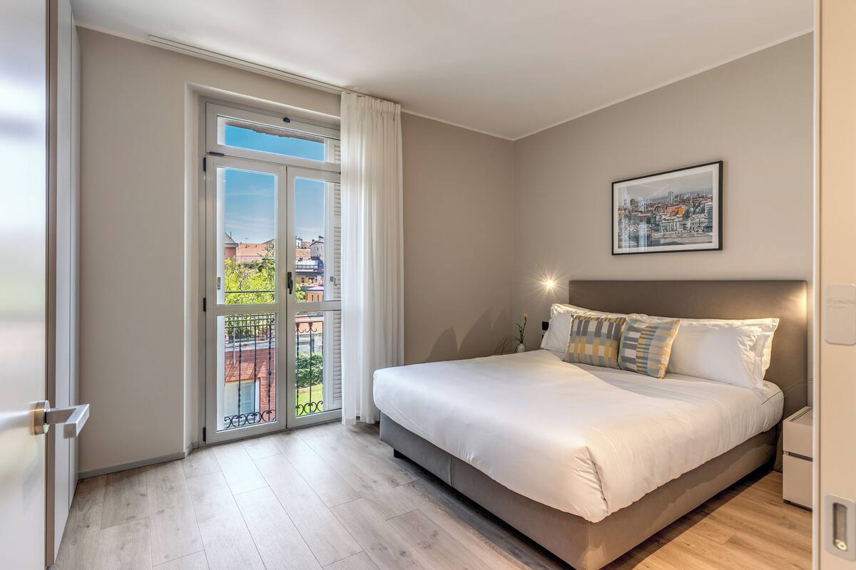 A bedroom with a large bed, a sliding glass door to a balcony, and a cityscape view.