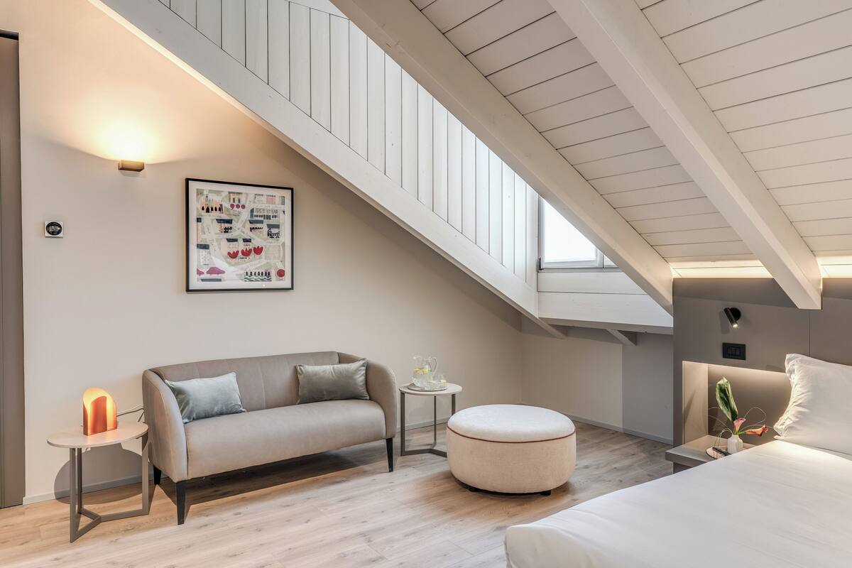 A modern loft bedroom with a sloped ceiling, a sofa, a round ottoman, a bedside table with a lamp, and a framed map on the wall.