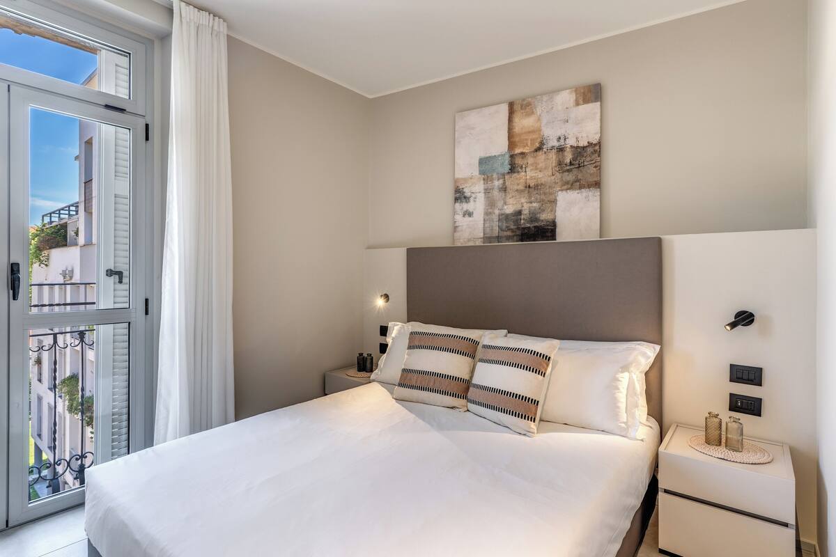 A neatly made bed with white linens and striped pillows, a bedside table with a lamp, and a view of the outside balcony.