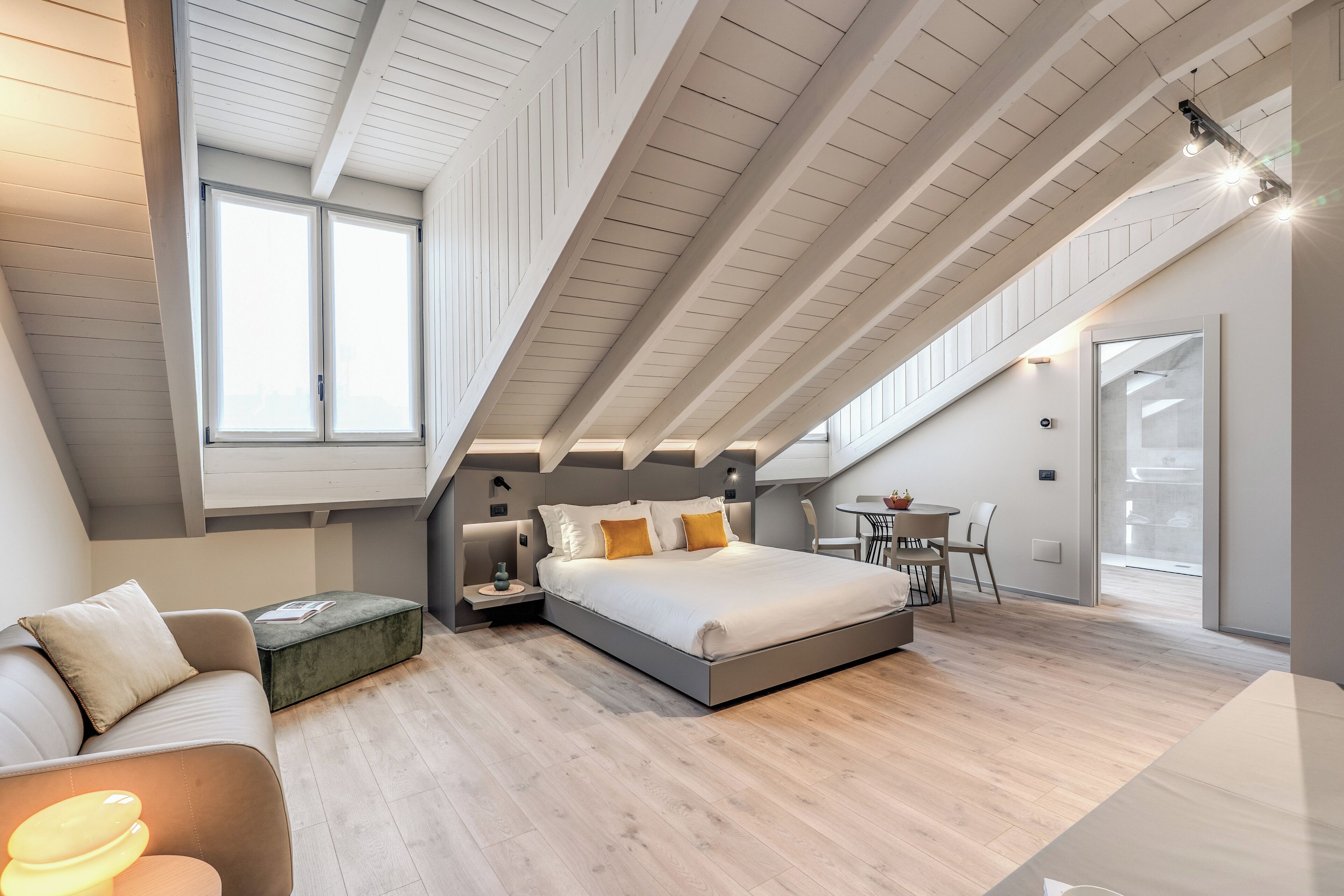 A modern loft bedroom with a large bed, a sofa, a dining table, and a staircase leading to an upper level.