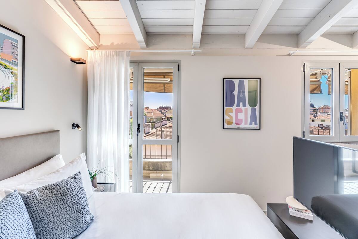 A modern hotel room with a large bed, a TV, a balcony with a view of buildings, and a framed artwork on the wall.
