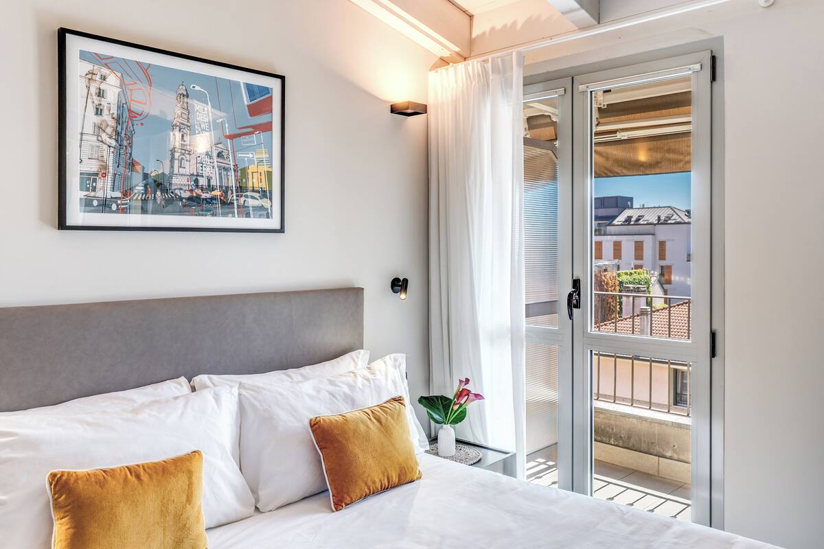 A hotel room with a bed, white bedding, a framed cityscape artwork, a nightstand with a vase of flowers, and a sliding glass door to a balcony.