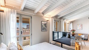 Habitación de hotel moderna con una cama grande, televisor, zona de comedor con mesa y sillas, y balcón con cortinas.