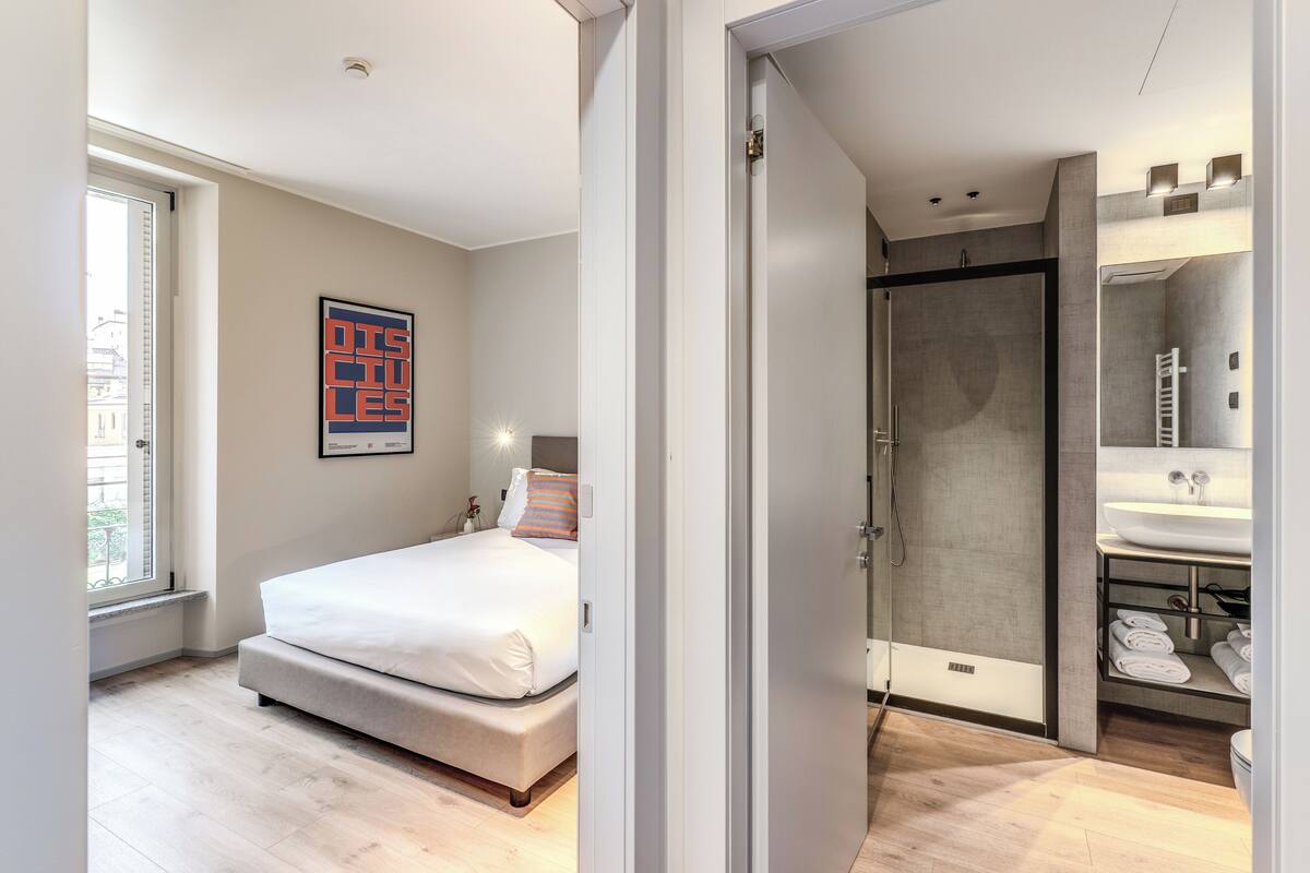 A modern bedroom with a bed, a shower, and a bathroom with a sink and towel rack.