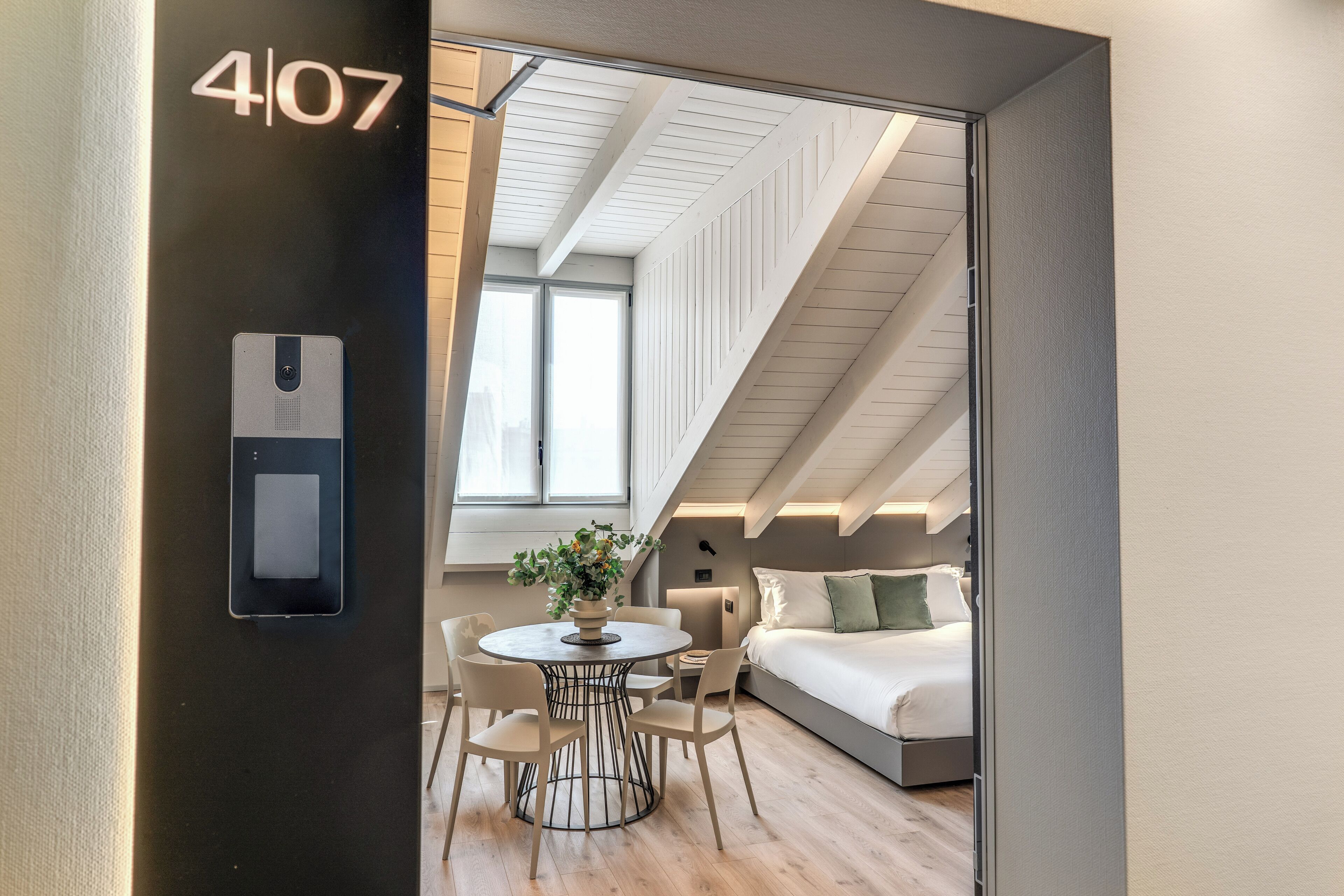 A modern hotel room with a lofted ceiling, a bed, a dining table with chairs, and a small balcony.