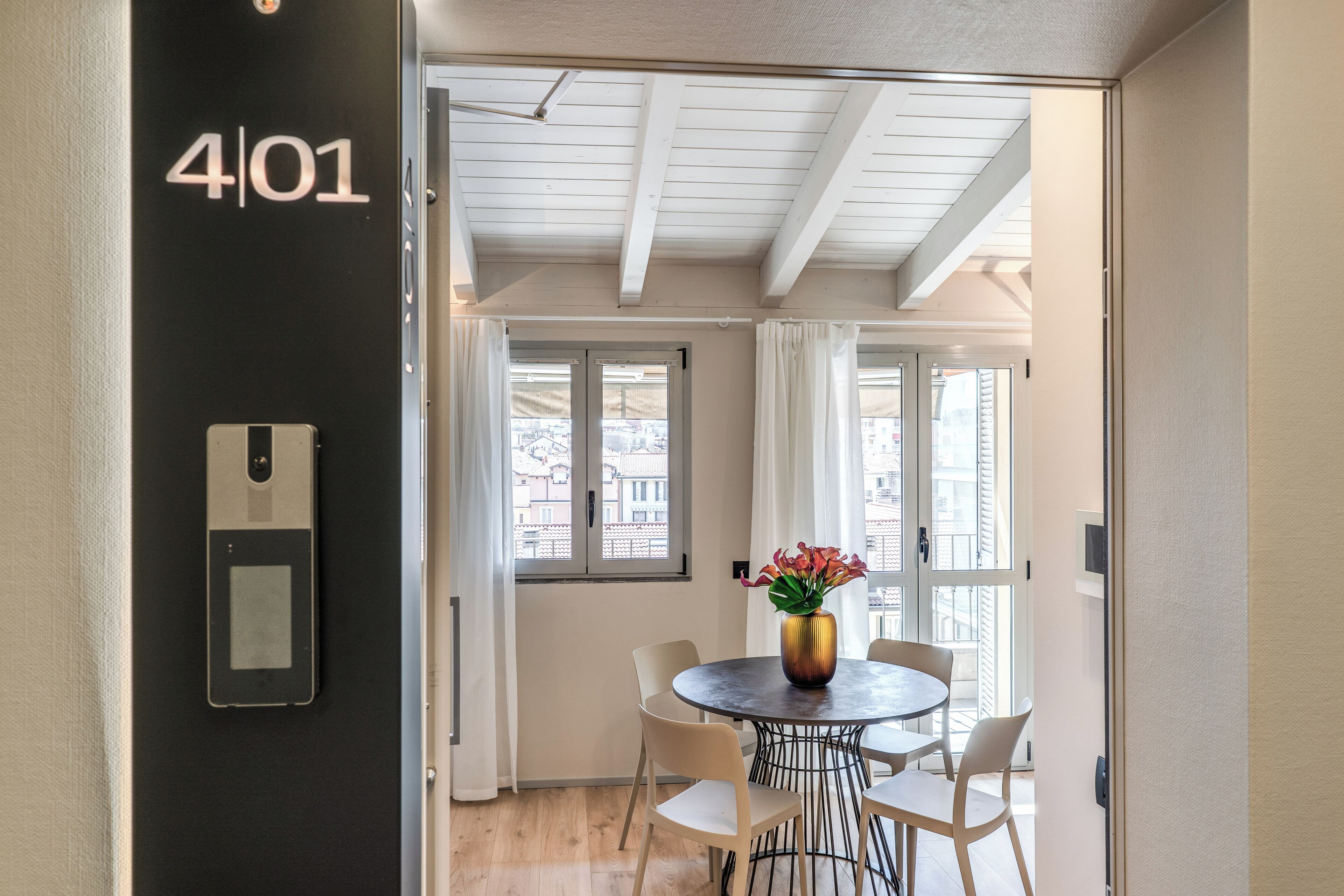 Habitación de hotel con una puerta negra que indica “4/01”, una mesa redonda de comedor con sillas blancas, un jarrón con flores y vistas a través de la ventana.