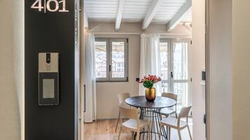 Habitación de hotel con una puerta negra que indica “4/01”, una mesa redonda de comedor con sillas blancas, un jarrón con flores y vistas a través de la ventana.