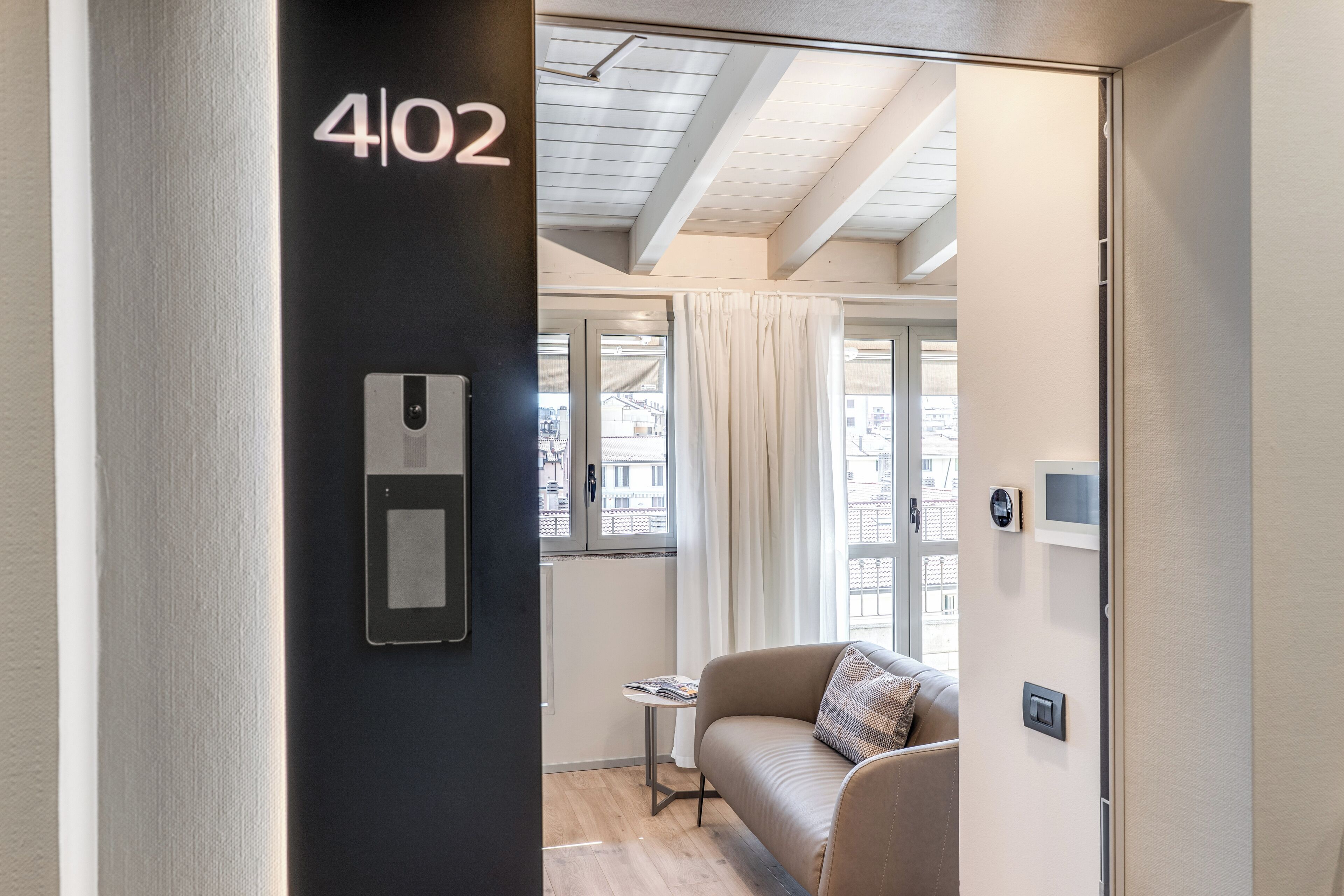 Une chambre d’hôtel moderne avec un mur noir sur lequel est affiché le numéro 402, une fenêtre avec des rideaux, un canapé beige et une petite table avec un magazine.