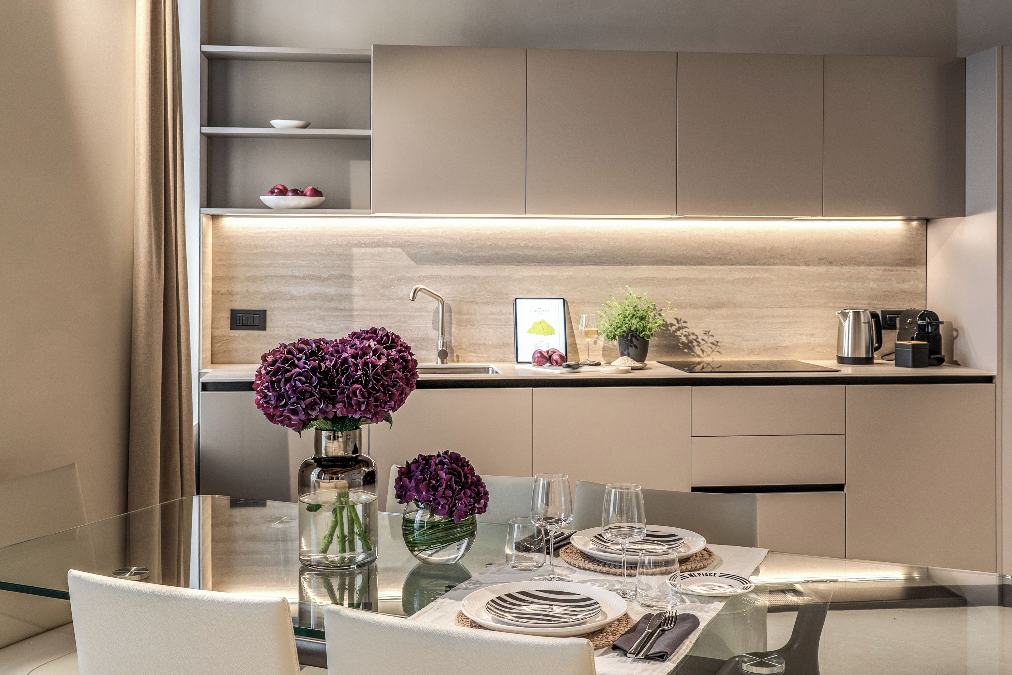 Eine moderne Küche mit einem Glastisch, zwei Vasen mit violetten Blumen und einer Kaffeemaschine.