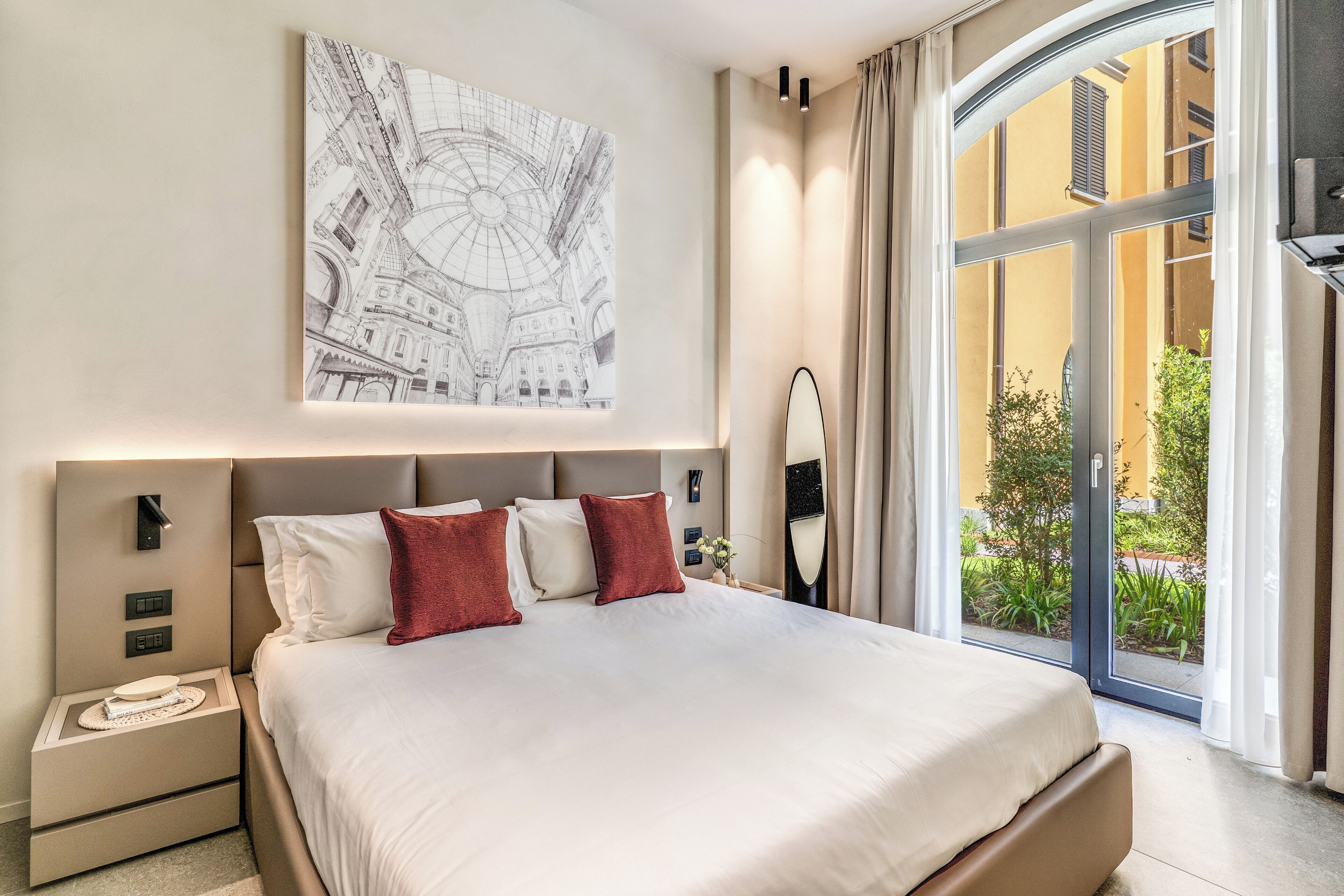 A modern hotel room with a large bed, a nightstand, a wall-mounted artwork, and a view of the outdoors.