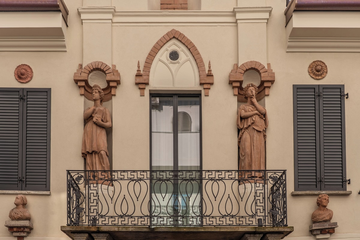 Fachada de edificio con dos estatuas en el balcón, una ventana con contraventanas y elementos decorativos sobre la ventana.