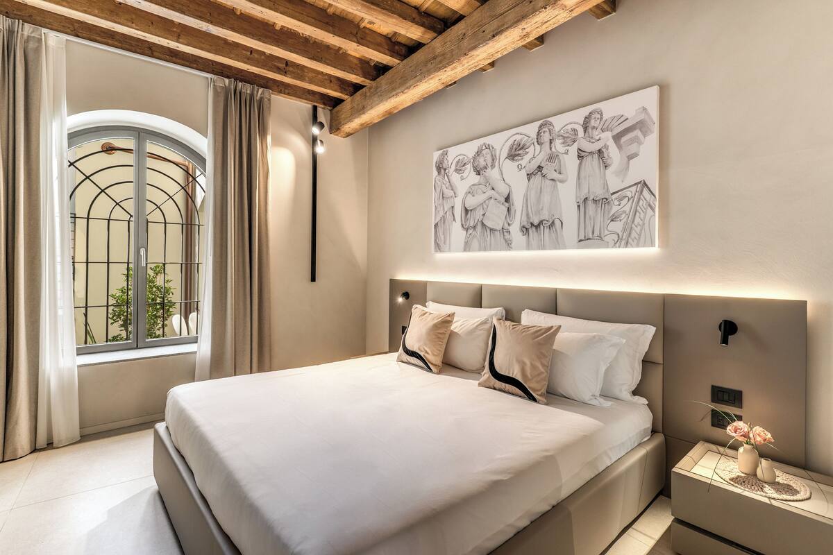 Un dormitorio con una cama grande, cabecera con iluminación integrada, ventana con cortinas y una obra de arte colgada en la pared.