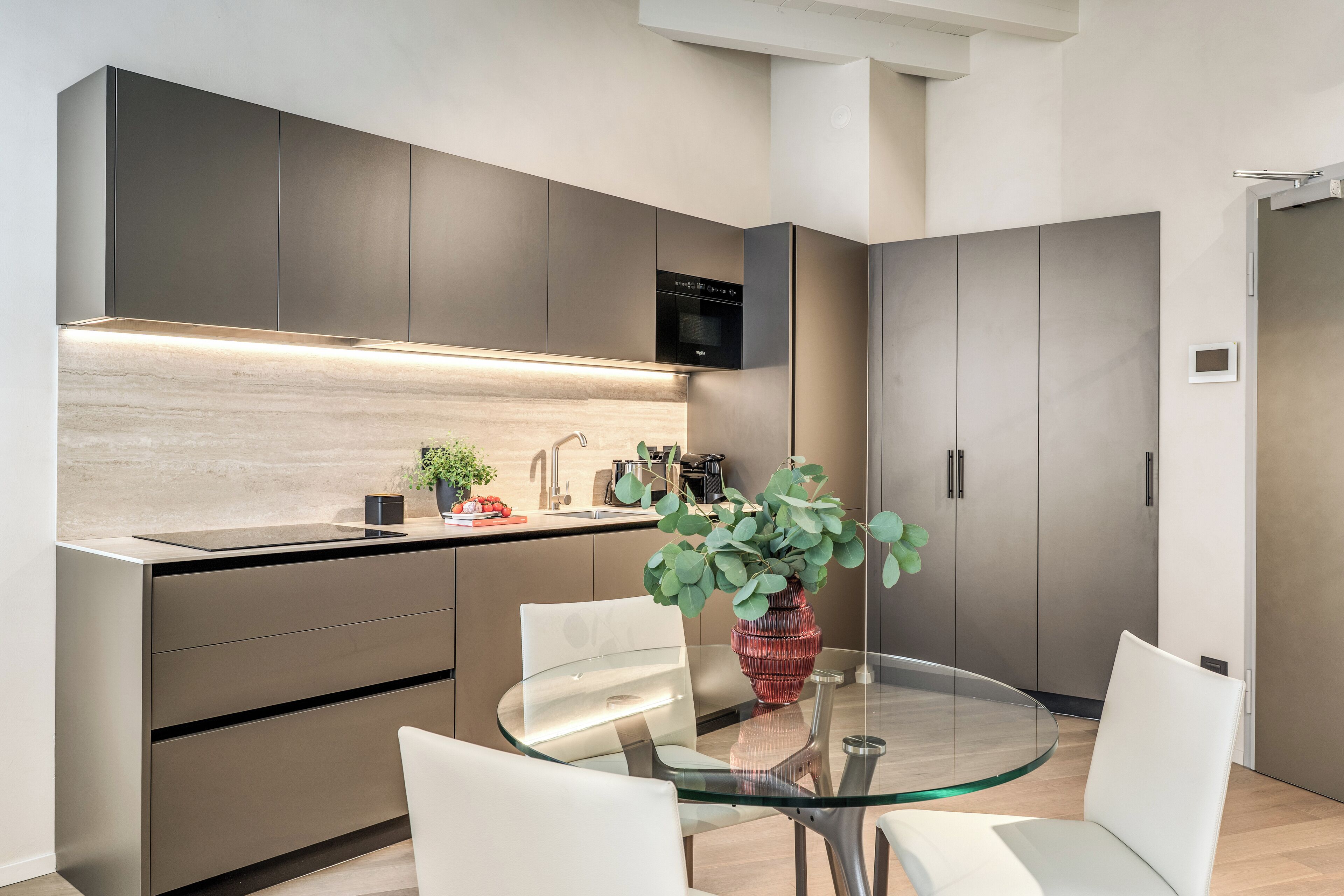 A modern kitchen with a glass dining table, stainless steel appliances, and a potted plant.