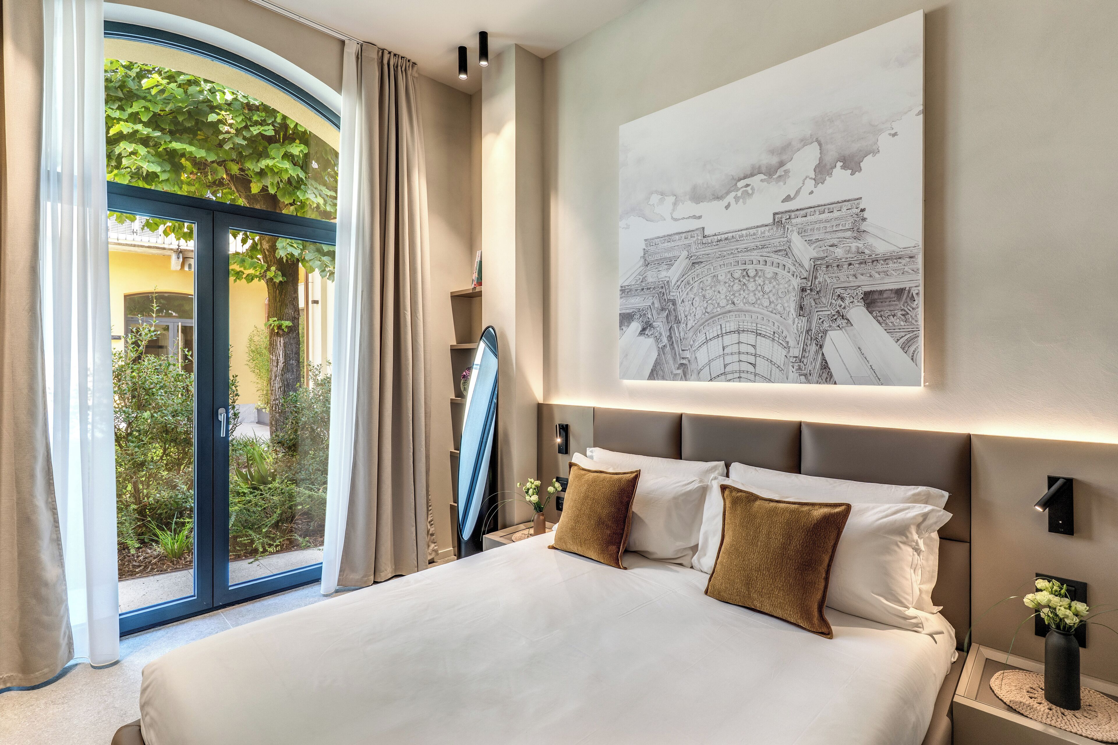 Quarto de hotel com uma cama grande, vista para o verde através de uma porta de vidro, uma obra de arte na parede representando um edifício e uma mesinha de cabeceira com um vaso de flores.