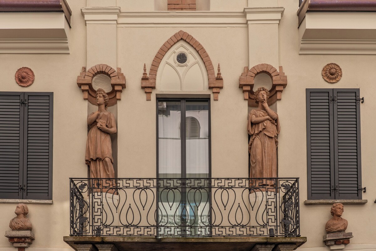 Fachada de edificio con dos estatuas en el balcón, una ventana con contraventanas y elementos decorativos sobre la ventana.