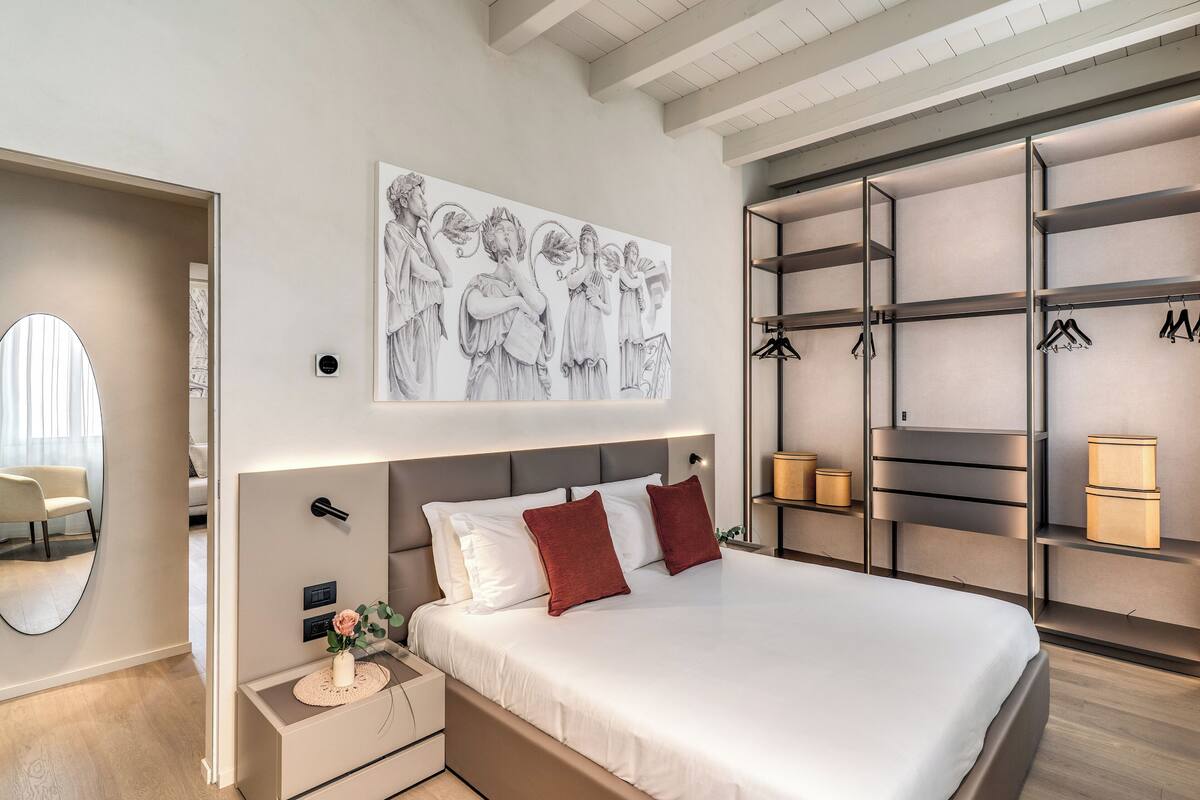 Un dormitorio moderno con una cama grande, cabecera con iluminación integrada, una obra de arte en la pared y un vestidor con estantes y espacio para colgar.
